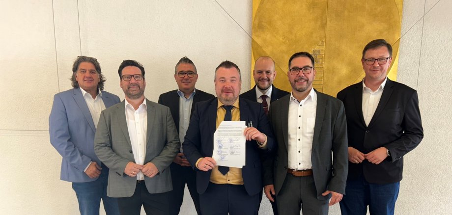 v.l.n.r.: Peter Spitzer, Holger Zwick, Björn Bernhard, Patrick Weißler, Felix Leidecker, Klaus Weber, Patrick Sema Sieben Männer in Anzügen stehen vor einer Wand, der mittlere Mann hält ein unterschriebenes Dokument in die Kamera